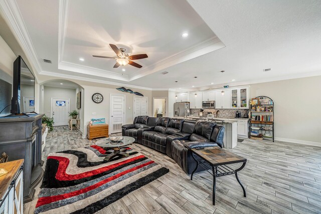 living room with ceiling fan, light wood-type flooring, a raised ceiling, and crown molding