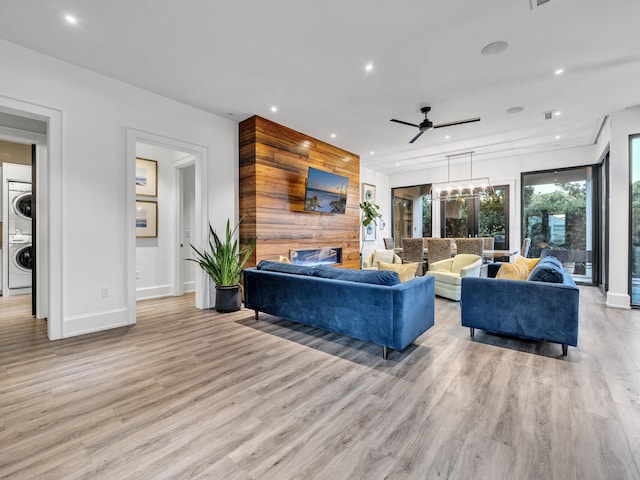 living room featuring light wood-type flooring, ceiling fan with notable chandelier, stacked washing maching and dryer, and a fireplace