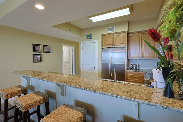 kitchen featuring kitchen peninsula, stainless steel fridge, backsplash, a kitchen breakfast bar, and light stone counters