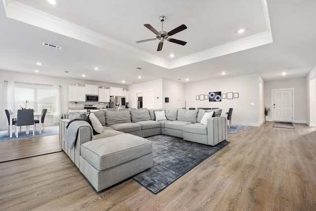 living room featuring ceiling fan, a tray ceiling, light wood-type flooring, and ornamental molding