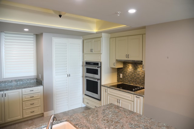 kitchen featuring cream cabinetry, black electric cooktop, double oven, dark stone countertops, and decorative backsplash