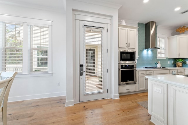 kitchen with white cabinets, ornamental molding, light hardwood / wood-style flooring, wall chimney range hood, and stainless steel appliances