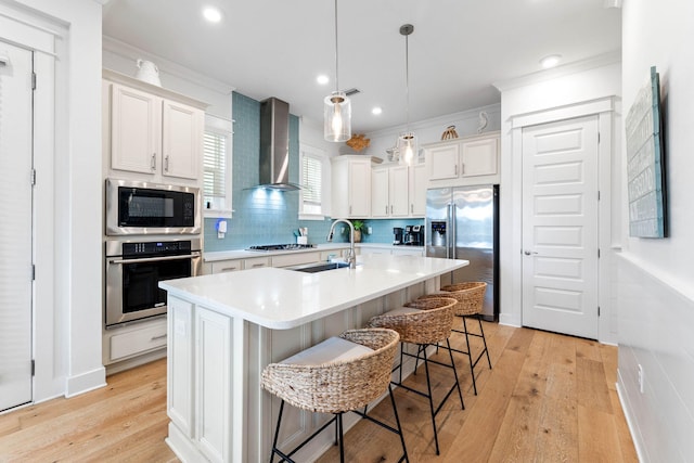 kitchen featuring appliances with stainless steel finishes, wall chimney exhaust hood, a kitchen island with sink, and light hardwood / wood-style floors