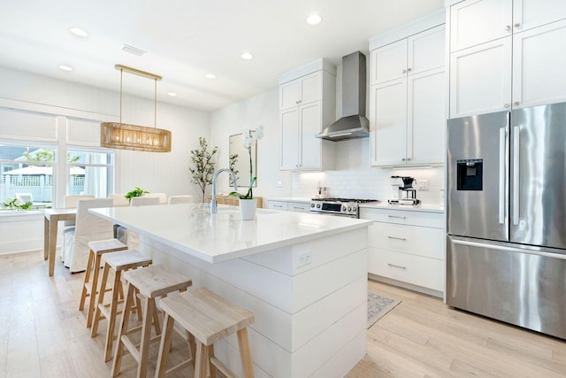 kitchen featuring light wood-type flooring, an island with sink, white cabinetry, wall chimney range hood, and appliances with stainless steel finishes