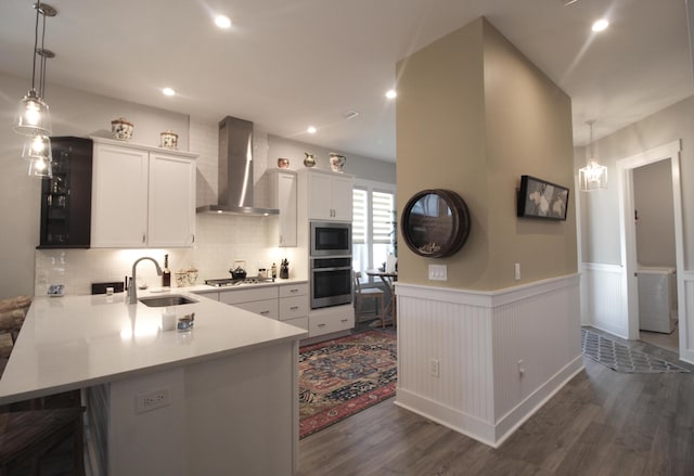 kitchen with sink, kitchen peninsula, wall chimney range hood, appliances with stainless steel finishes, and dark hardwood / wood-style flooring