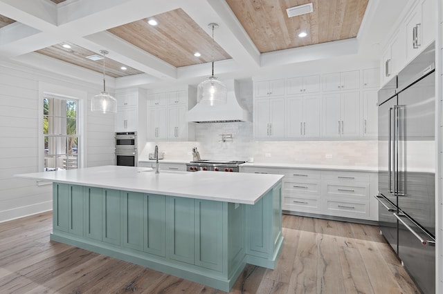 kitchen with appliances with stainless steel finishes, custom range hood, a large island with sink, white cabinetry, and light hardwood / wood-style flooring