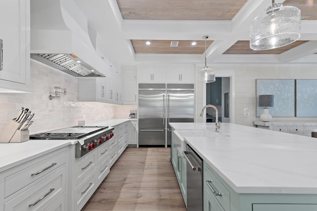 kitchen featuring an island with sink, white cabinetry, hanging light fixtures, wall chimney range hood, and appliances with stainless steel finishes