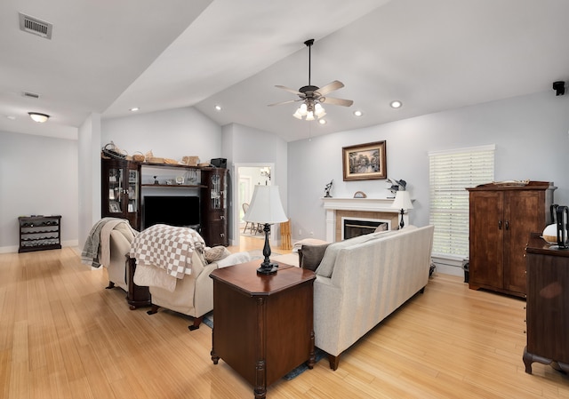 living room with ceiling fan, light wood-type flooring, a tiled fireplace, and vaulted ceiling