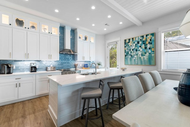 kitchen featuring white cabinetry, a kitchen bar, beam ceiling, a wealth of natural light, and wall chimney range hood