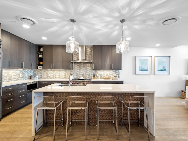 kitchen with a kitchen island with sink, wall chimney range hood, dark brown cabinets, and light hardwood / wood-style floors