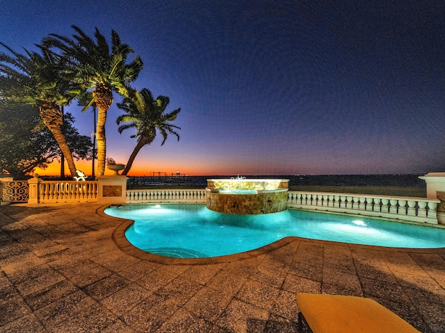 pool at dusk with an in ground hot tub, a water view, and a patio area