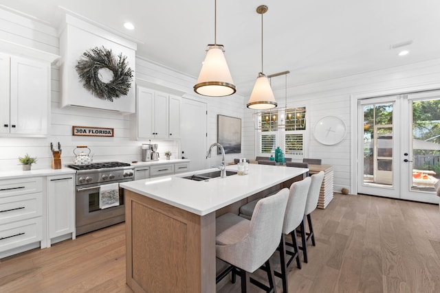 kitchen featuring light wood-type flooring, high end stainless steel range oven, an island with sink, white cabinetry, and decorative light fixtures