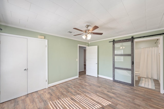 unfurnished bedroom featuring ceiling fan, hardwood / wood-style floors, crown molding, a barn door, and ensuite bathroom