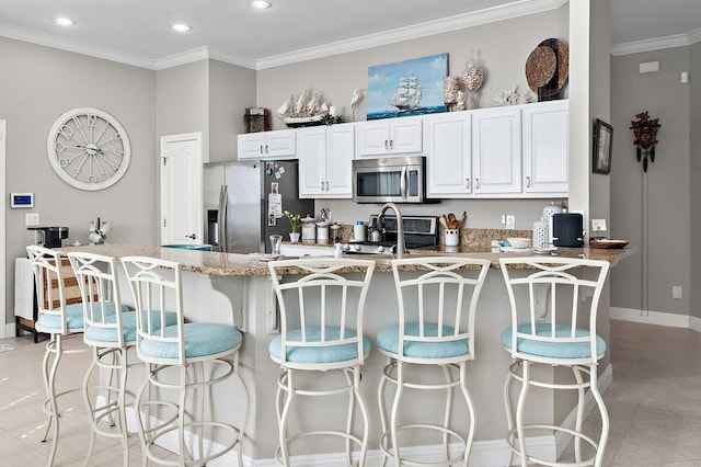 kitchen with light stone counters, ornamental molding, appliances with stainless steel finishes, a kitchen bar, and white cabinetry