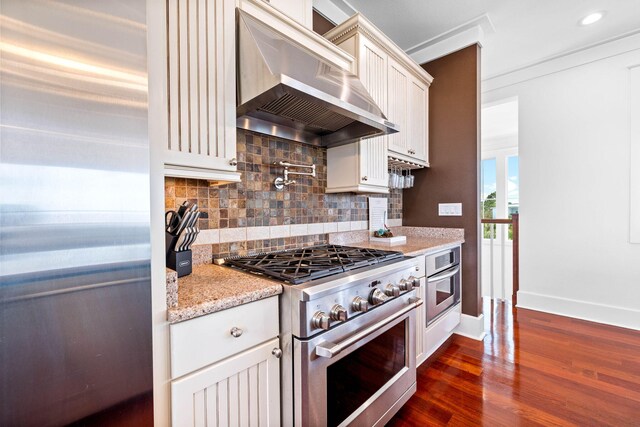 kitchen featuring wall chimney exhaust hood, tasteful backsplash, dark wood-type flooring, stainless steel appliances, and light stone countertops