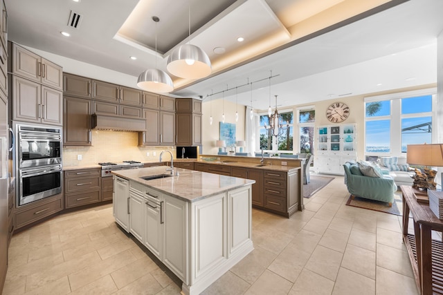 kitchen featuring pendant lighting, a kitchen island with sink, sink, a tray ceiling, and light stone counters