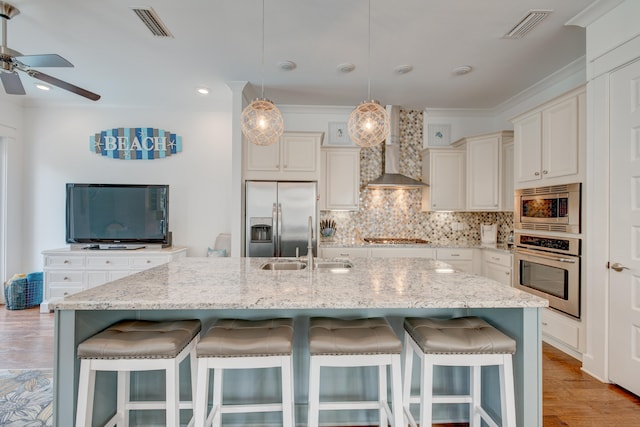 kitchen featuring an island with sink, light wood-type flooring, wall chimney exhaust hood, a breakfast bar area, and appliances with stainless steel finishes