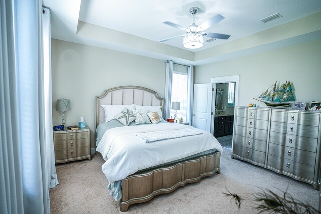 bedroom with ceiling fan, light colored carpet, a tray ceiling, and ensuite bath