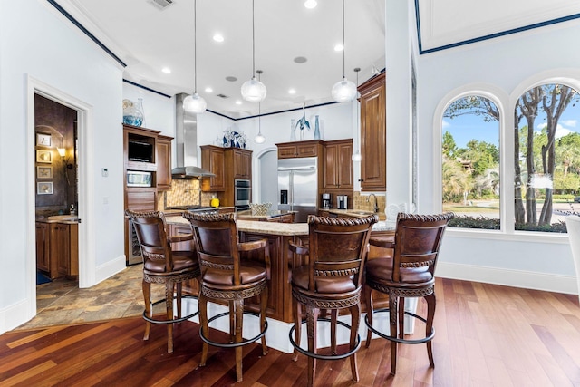 kitchen with wall chimney range hood, built in appliances, light stone counters, decorative backsplash, and dark hardwood / wood-style floors