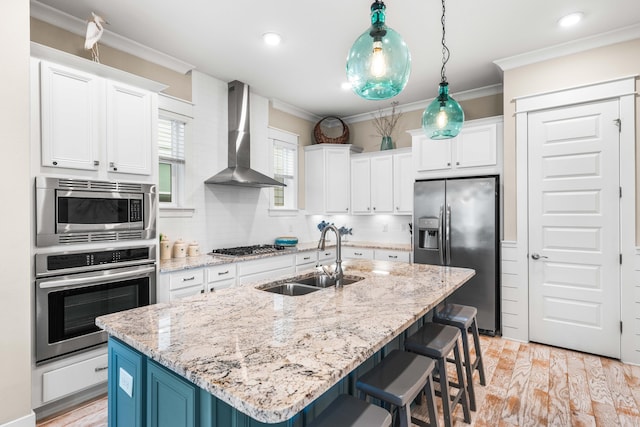 kitchen featuring wall chimney range hood, an island with sink, appliances with stainless steel finishes, pendant lighting, and sink