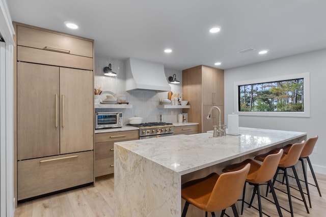 kitchen featuring custom exhaust hood, stainless steel range oven, paneled fridge, and a center island with sink