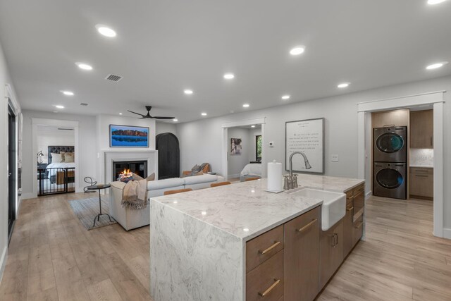 kitchen featuring light hardwood / wood-style floors, stacked washer and clothes dryer, sink, ceiling fan, and a kitchen island with sink