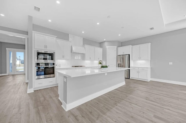 kitchen with white cabinets, a large island with sink, premium range hood, light hardwood / wood-style floors, and stainless steel appliances