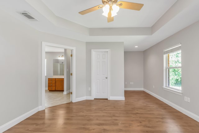 unfurnished bedroom with light wood-type flooring, connected bathroom, a tray ceiling, and ceiling fan