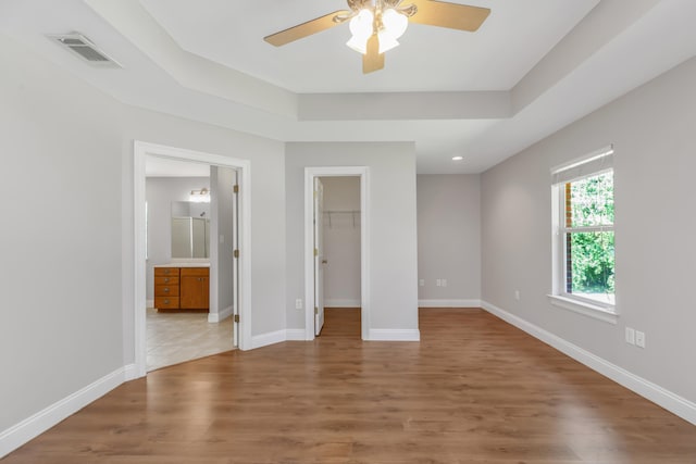 unfurnished bedroom with a spacious closet, ceiling fan, a raised ceiling, ensuite bathroom, and light wood-type flooring