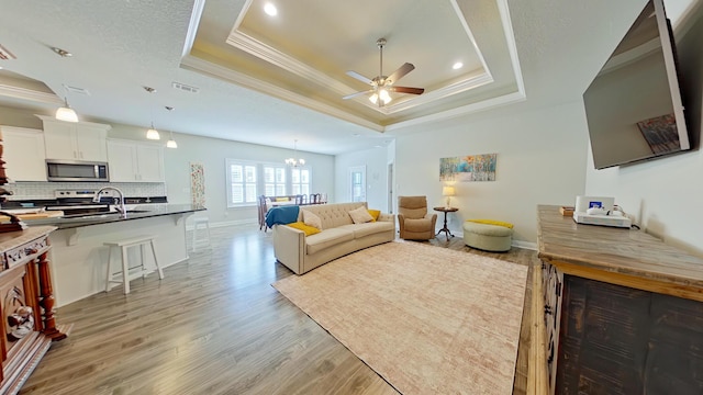 living room with ceiling fan with notable chandelier, sink, a raised ceiling, light wood-type flooring, and crown molding