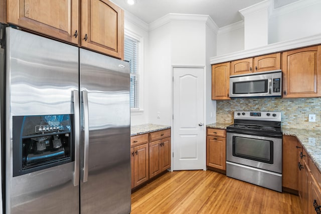 kitchen featuring stainless steel appliances, light stone counters, crown molding, decorative backsplash, and light wood-type flooring