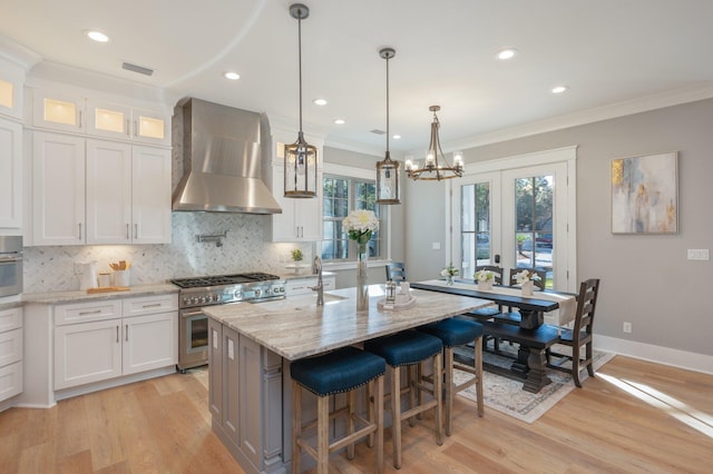 kitchen with french doors, wall chimney exhaust hood, stainless steel appliances, a center island with sink, and white cabinets