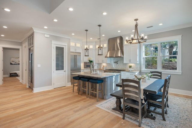 kitchen with white cabinets, decorative light fixtures, wall chimney exhaust hood, and an island with sink