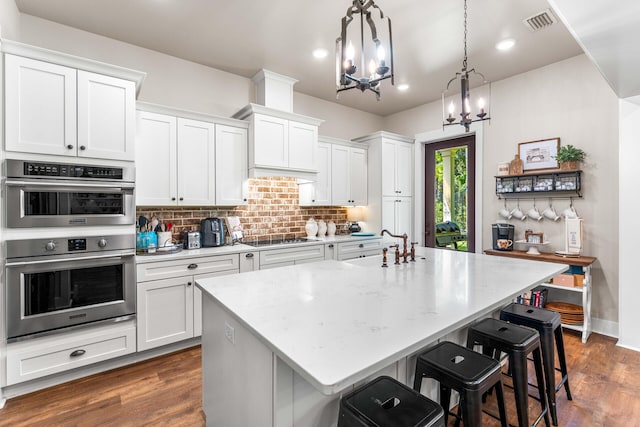 kitchen with a kitchen breakfast bar, white cabinetry, a kitchen island with sink, double oven, and decorative light fixtures