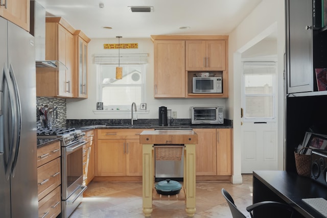 kitchen with wall chimney exhaust hood, stainless steel appliances, dark stone countertops, decorative light fixtures, and light brown cabinetry