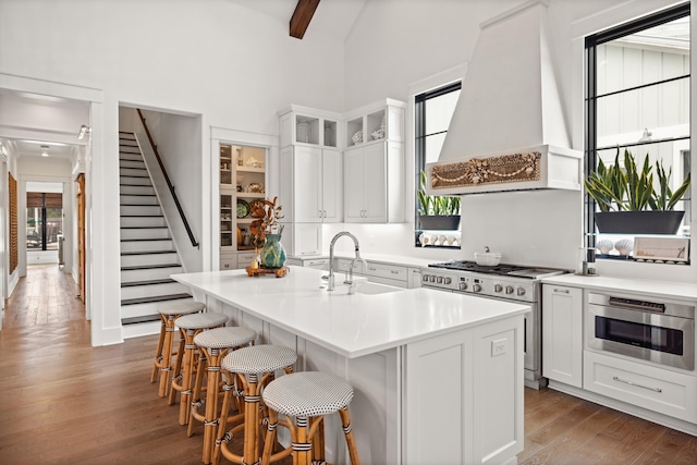 kitchen featuring custom range hood, stainless steel appliances, hardwood / wood-style flooring, a center island with sink, and white cabinetry