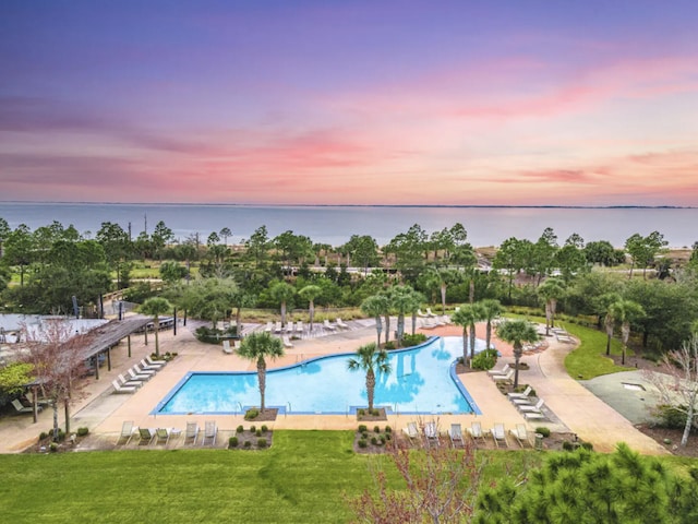 pool at dusk featuring a patio, a water view, and a lawn