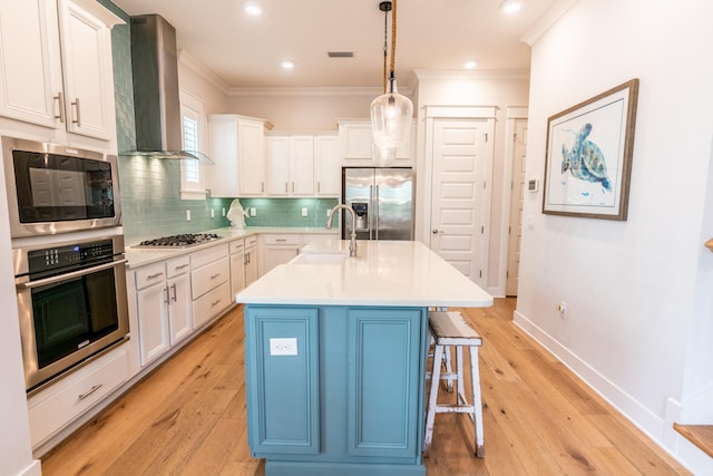 kitchen with wall chimney range hood, sink, appliances with stainless steel finishes, pendant lighting, and a center island with sink