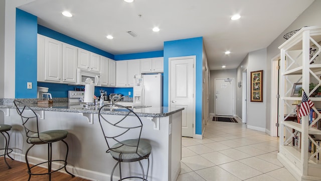 kitchen featuring kitchen peninsula, white cabinets, a breakfast bar, dark stone counters, and white appliances