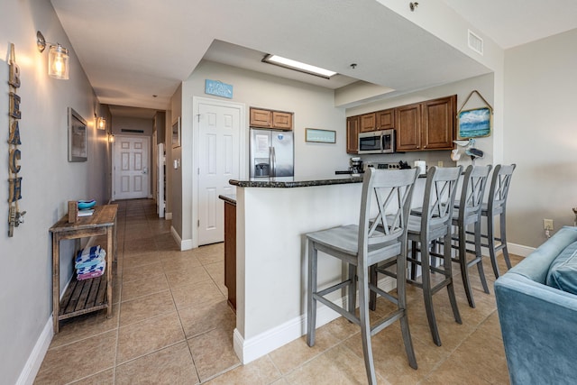 kitchen featuring appliances with stainless steel finishes, a breakfast bar, kitchen peninsula, dark stone counters, and light tile patterned floors