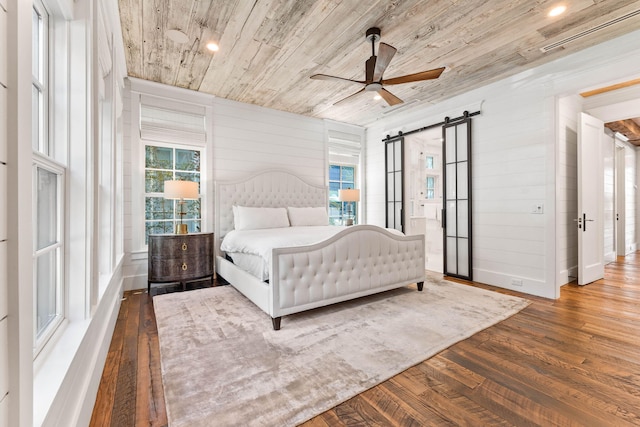 bedroom with dark wood-type flooring, wooden ceiling, a barn door, ceiling fan, and wood walls