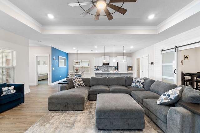 living room with ceiling fan, a textured ceiling, a barn door, a tray ceiling, and light wood-type flooring