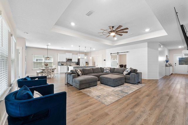 living room featuring light hardwood / wood-style flooring, ornamental molding, a barn door, a raised ceiling, and ceiling fan