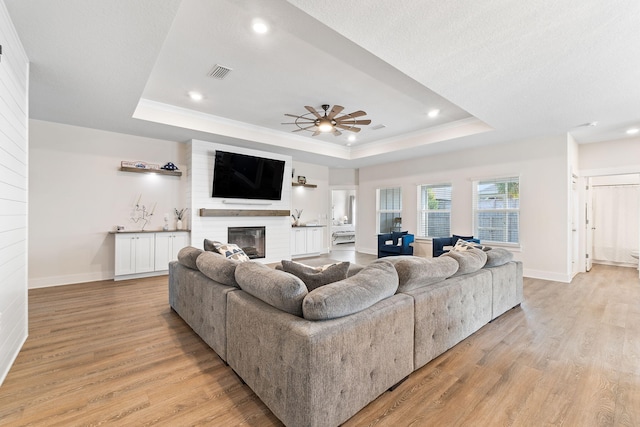 living room featuring a large fireplace, a textured ceiling, a tray ceiling, light hardwood / wood-style floors, and ceiling fan