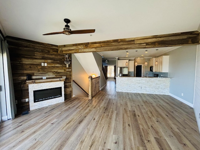 unfurnished living room featuring beamed ceiling, light wood finished floors, a glass covered fireplace, a ceiling fan, and baseboards
