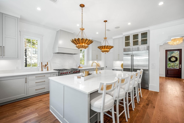 kitchen with stove, built in refrigerator, an island with sink, hardwood / wood-style flooring, and custom exhaust hood