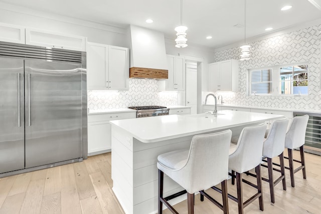 kitchen with premium range hood, white cabinetry, a kitchen island with sink, decorative light fixtures, and stainless steel appliances