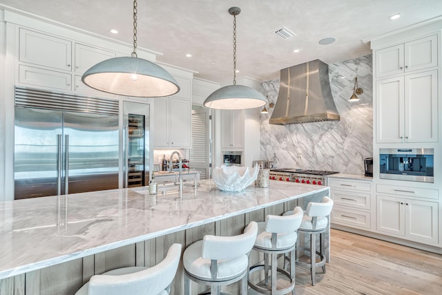 kitchen featuring built in refrigerator, pendant lighting, light stone counters, and wall chimney exhaust hood