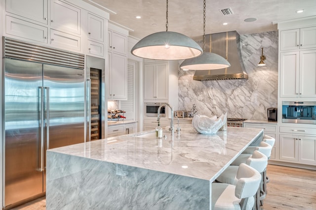 kitchen with wall chimney range hood, sink, stainless steel appliances, decorative light fixtures, and a large island with sink