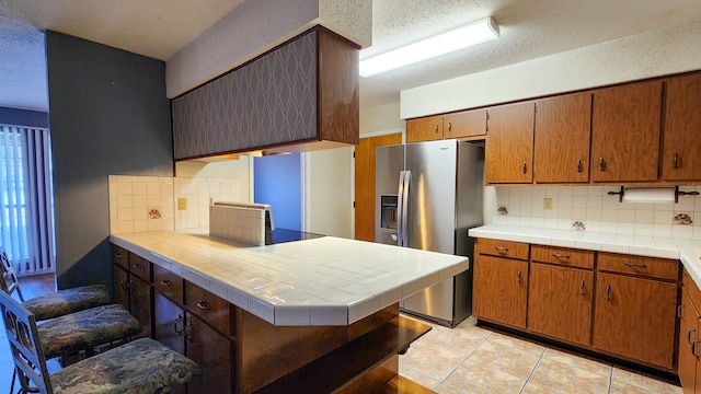 kitchen featuring backsplash, kitchen peninsula, tile counters, light tile patterned floors, and stainless steel refrigerator with ice dispenser